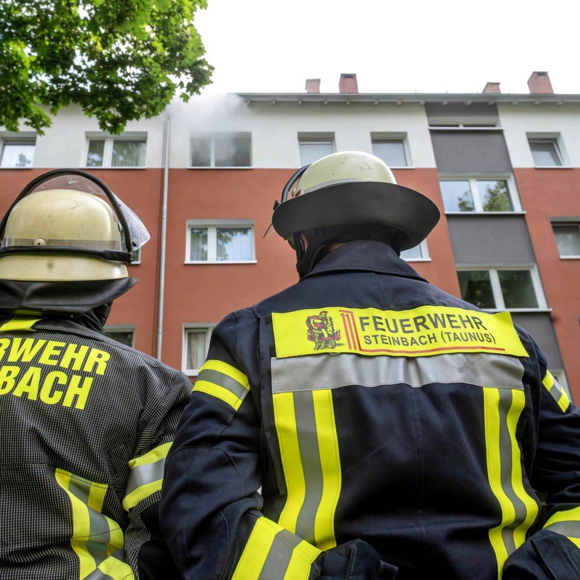 Zwei Feuerwehrleute schauen auf ein Mehrfamilienhaus, aus dessen oberstem Stockwerk Rauch herausquillt