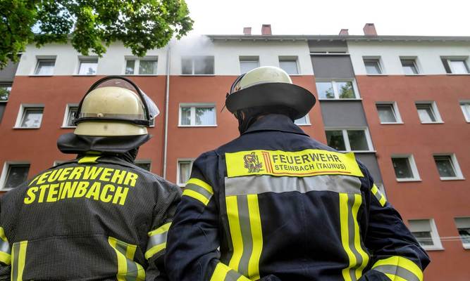 Zwei Feuerwehrleute schauen auf ein Mehrfamilienhaus, aus dessen oberstem Stockwerk Rauch herausquillt