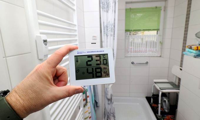 Eine Hand hält ein Thermometer mit Hygrometer-Funktion in einem Badezimmer fest und misst Raumtemperatur und Luftfeuchtigkeit.