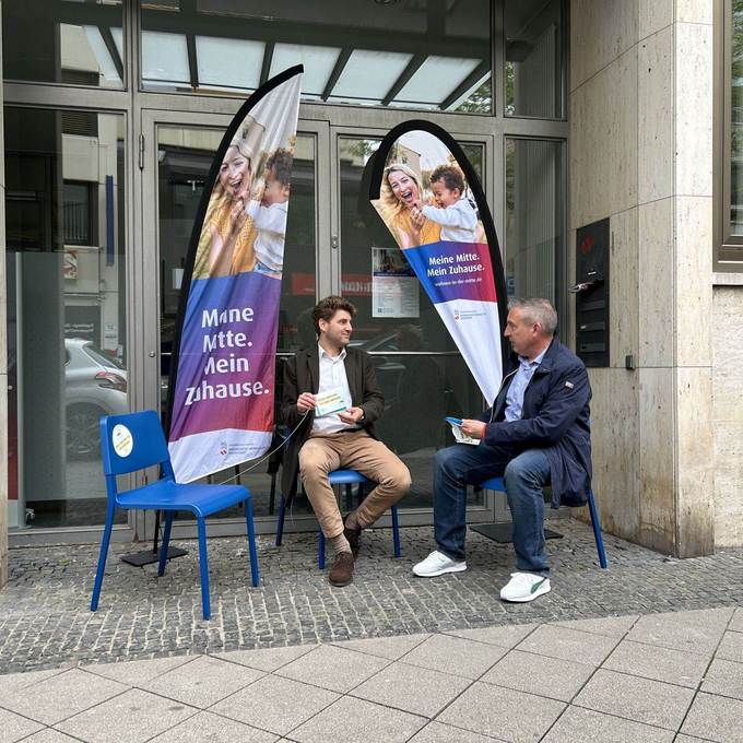 Zwei Männer sitzen auf Stühlen vor einem Bürogebäude und sprechen miteinander. 