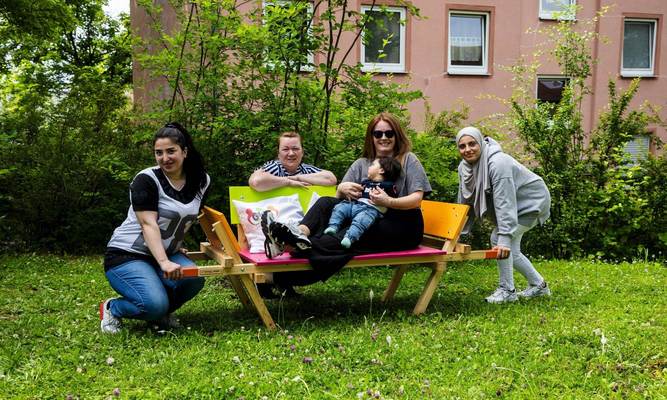 Eine Frau und ein kleines Kind sitzen auf einer bunt bemalten Bank, drei weitere Frauen hocken links, rechts und hinter der Bank