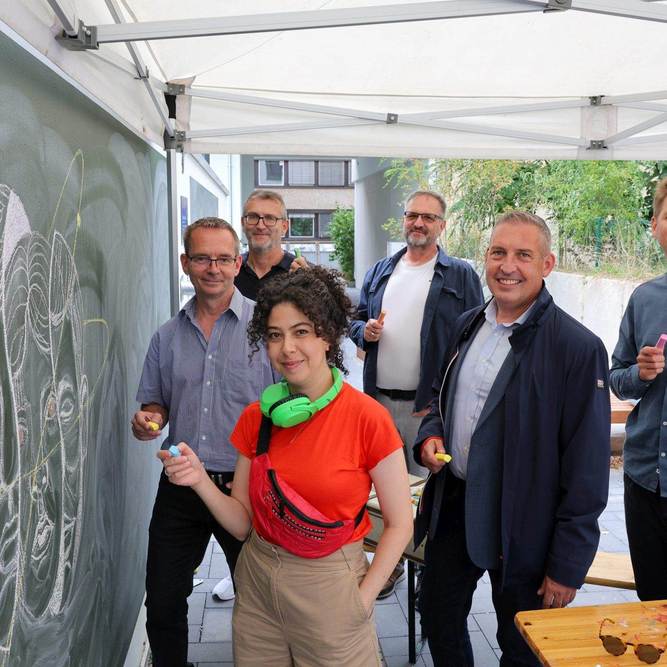 Gruppenfoto vor der Tafel: Künstlerin Aliaa Abou Khaddour sowie (v. li.) Matthias Otto (Leiter Servicecenter Kassel), Frank Jungermann (Kundenmanager), Friedemann Bunge (Projektleiter Neubau), Sascha Holstein (Leiter Regionalcenter Kassel) und Alexander Hauschild (Projektleiter Soziale Quartiersentwicklung).