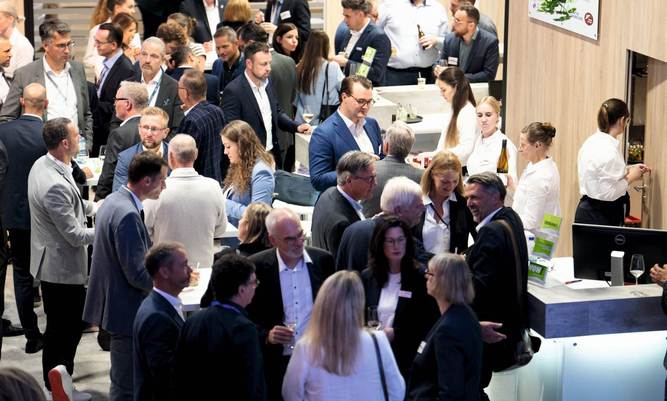 Viele Menschen, alle schick gekleidet, stehen an einem Messestand und unterhalten sich miteinander