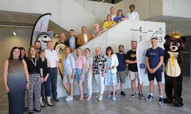 Gruppenfoto mit rund 25 Personen, zwei Maskottchen und zwei Beachflags in einem Treppenhaus, darunter Sascha Holstein, NHW-Regionalcenterleiter Kassel, Alexander Hauschild (Soziale Quartiersentwicklung NHW) und Kassels Sportdezernent Heiko Lehmkuhl