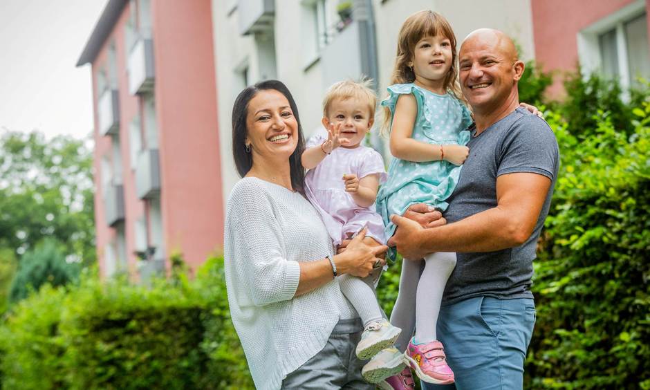 Familie steht vor einem Haus. Die Eltern haben die Kinder auf dem Arm.