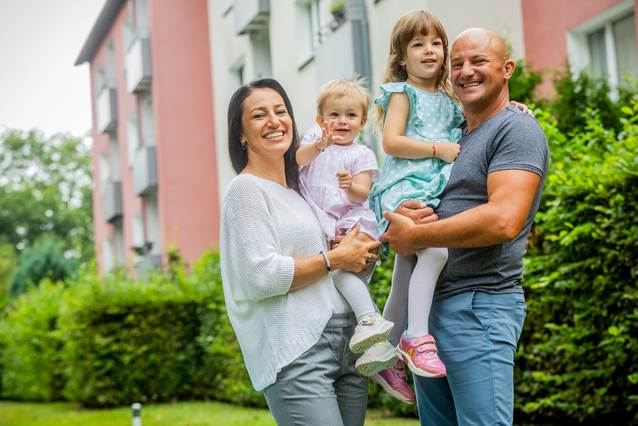 Familie steht vor einem Haus. Die Eltern haben die Kinder auf dem Arm.