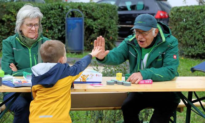 Ein älterer Mann und eine ältere Dame sitzen hinter einem Holztisch, der Mann gibt einem Jungen High Five