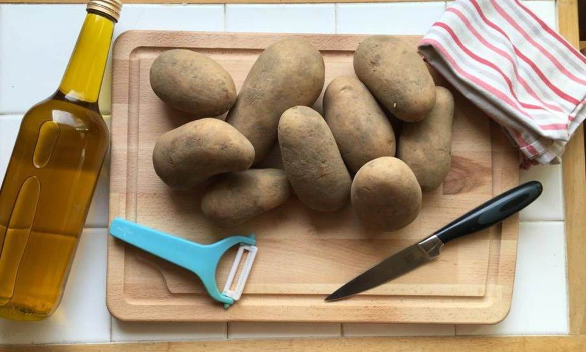 Kartoffeln, Messer, Schäler und Tuch liegen auf einem Holzbrett. Daneben liegt eine Flasche Öl.