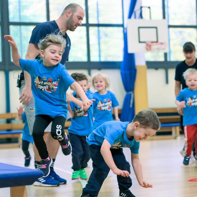 Kinder in blauen T-Shirts mit der Aufschrift "Sport macht Spaß" spielen und toben in einer Sporthalle.