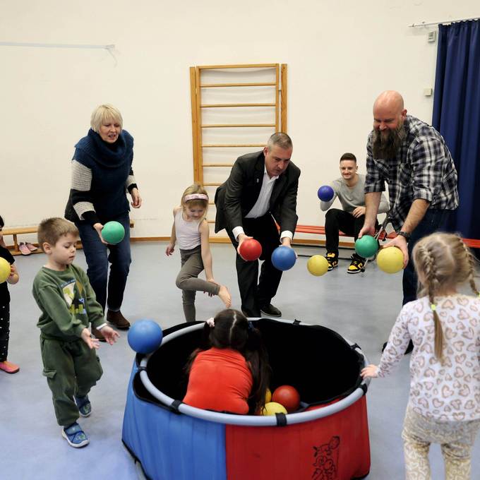 Mehrere Erwachsene spielen mit Kindern in einer Turnhalle. In der Mitte steht eine Art Bällebad, in dem auch Kinder sitzen. Alle werfen sich gegenseitig Bälle zu.