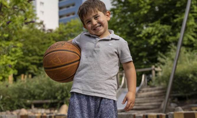 Ein kleiner Junge mit einem Basketball in der Hand steht auf einem Spielplatz vor einem Wohnhochhaus.