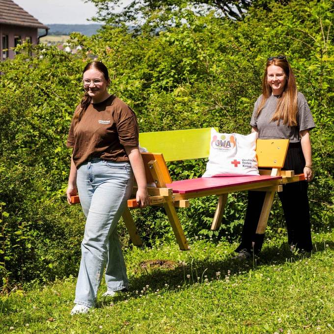 Zwei Frauen tragen eine bunt bemalte Holzbank über eine Rasenfläche. Im Hintergrund Büsche und ein Gebäude.