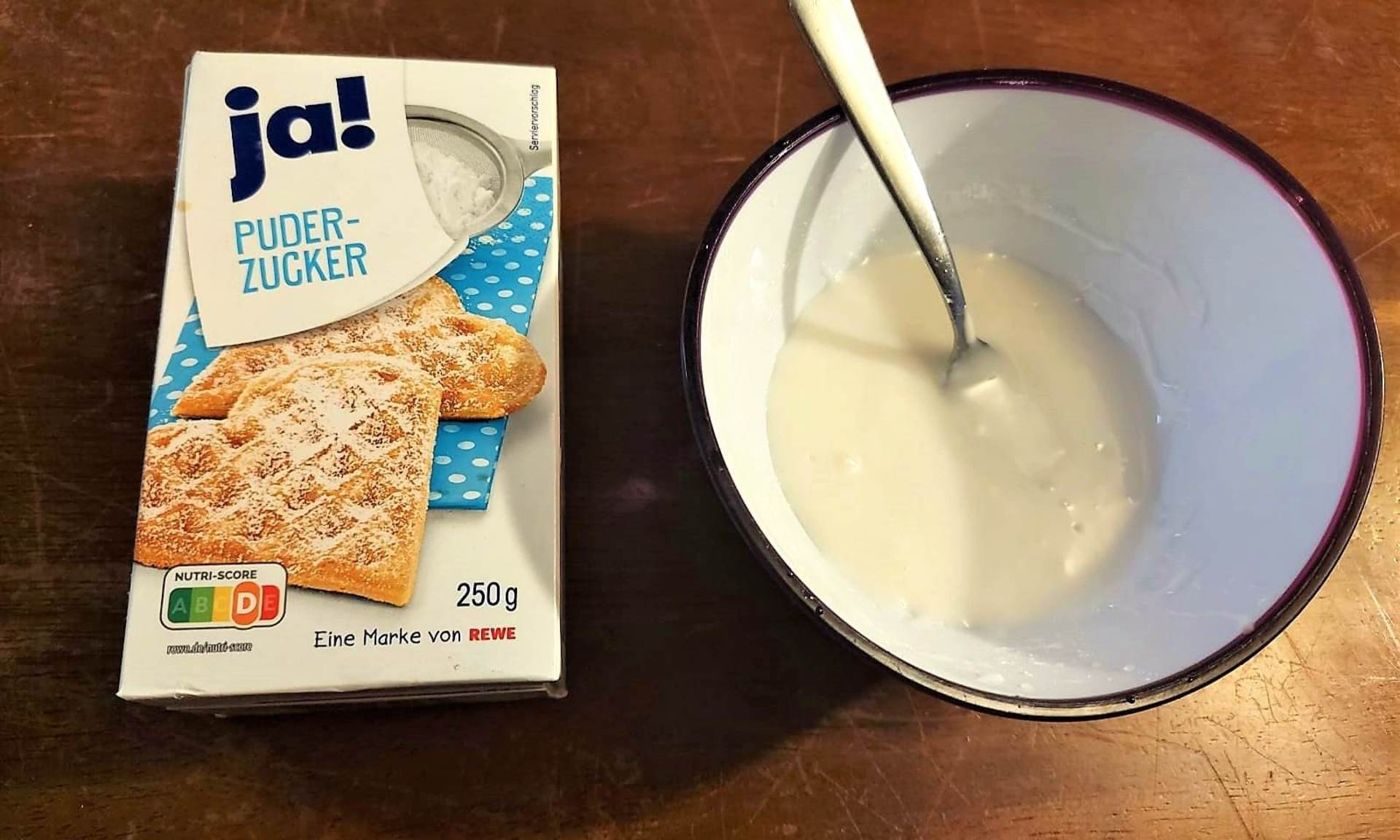 Zuckerguss in kleiner Schüssel mit Löffel. Daneben liegt ein Päckchen Puderzucker.