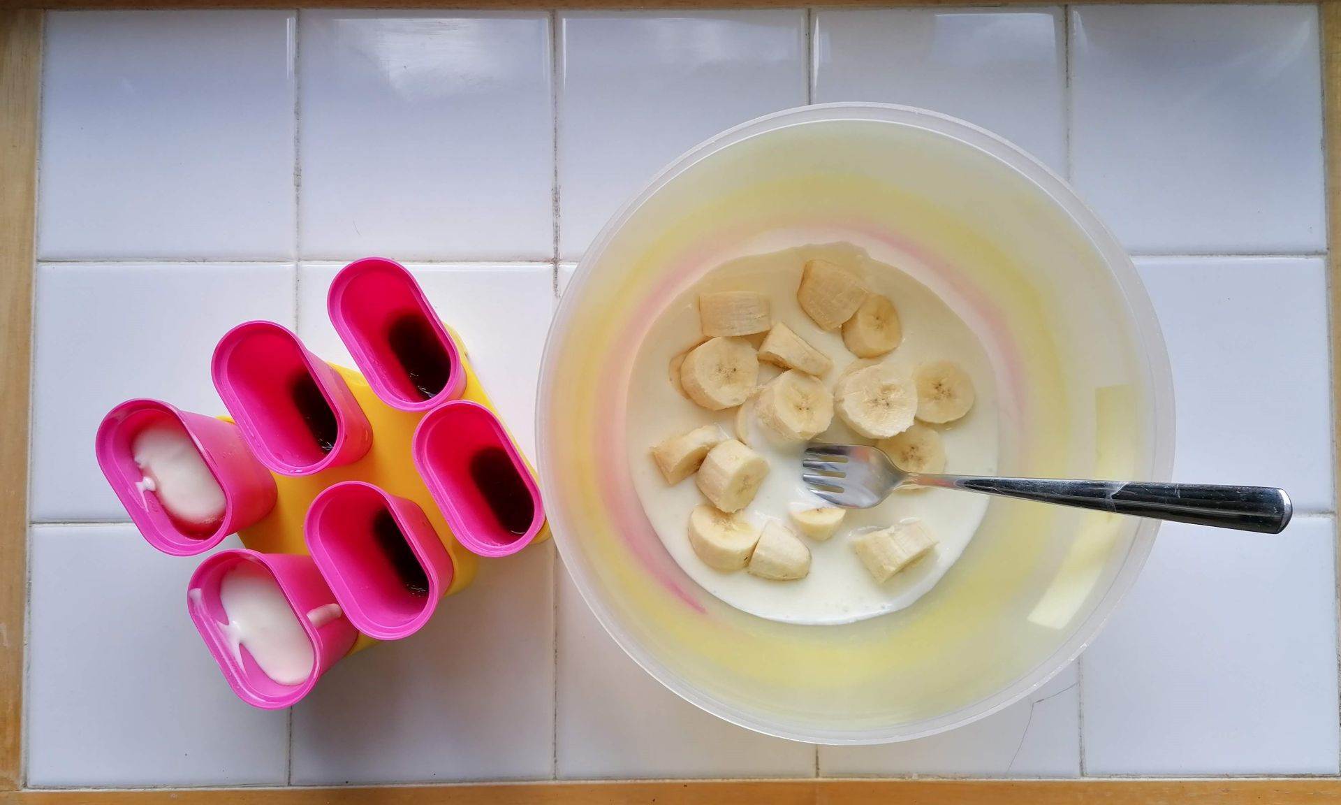 Bananenstücke und Joghurt in einer Schüssel mit Löffel. Daneben steht ein Eiszubereiter für Eis am Stil.