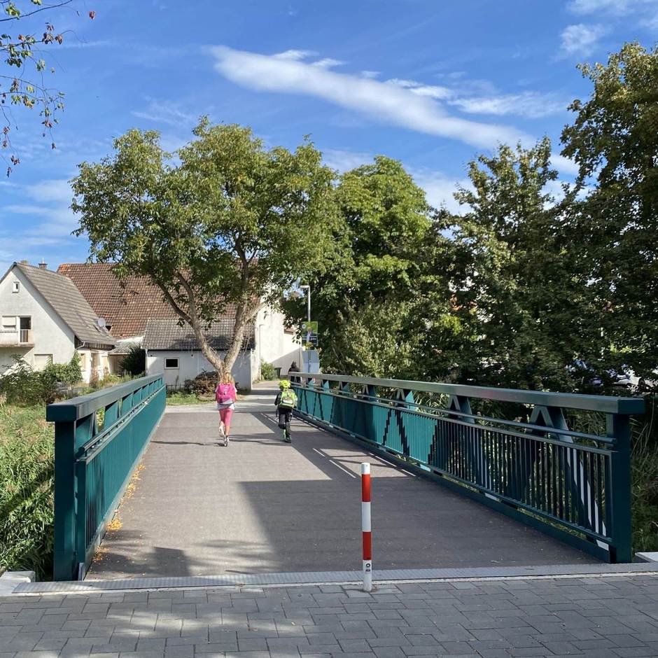 Ein Schulsteg der einen kleinen Fluss überquert. Kinder laufen über ihn hinüber. Grüne Vegetation ummantelt das Bild