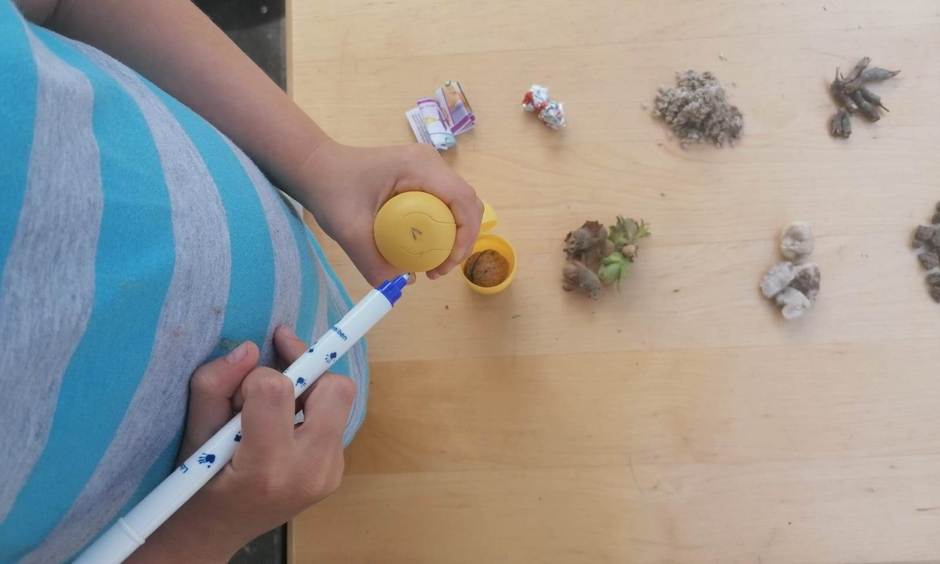 Junge beschriftet eine kleine, gelbe Verpackung. Auf dem Tisch liegen kleine Mengen von Reis, Nudeln, Erbsen, Steine etc.