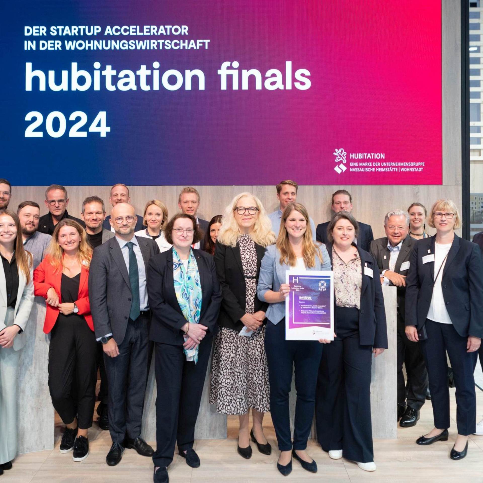 Gruppenfoto mit vielen gut gelaunten und lachenden Menschen, darüber ein Screen mit der Aufschrift "hubitation finals 2024"